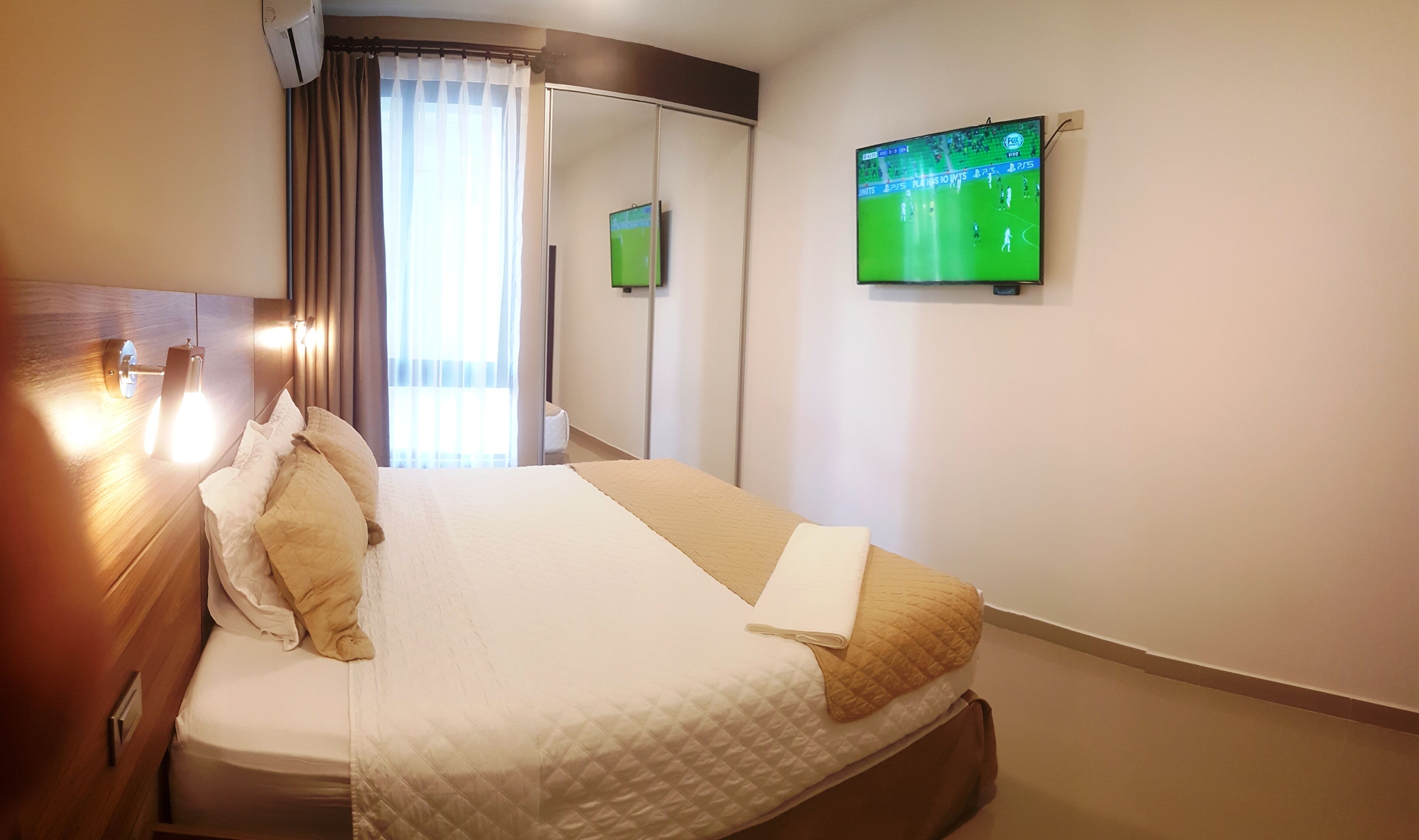 Habitación de hotel con una cama, televisión montada en la pared, ventana con cortinas y aire acondicionado también en la pared.