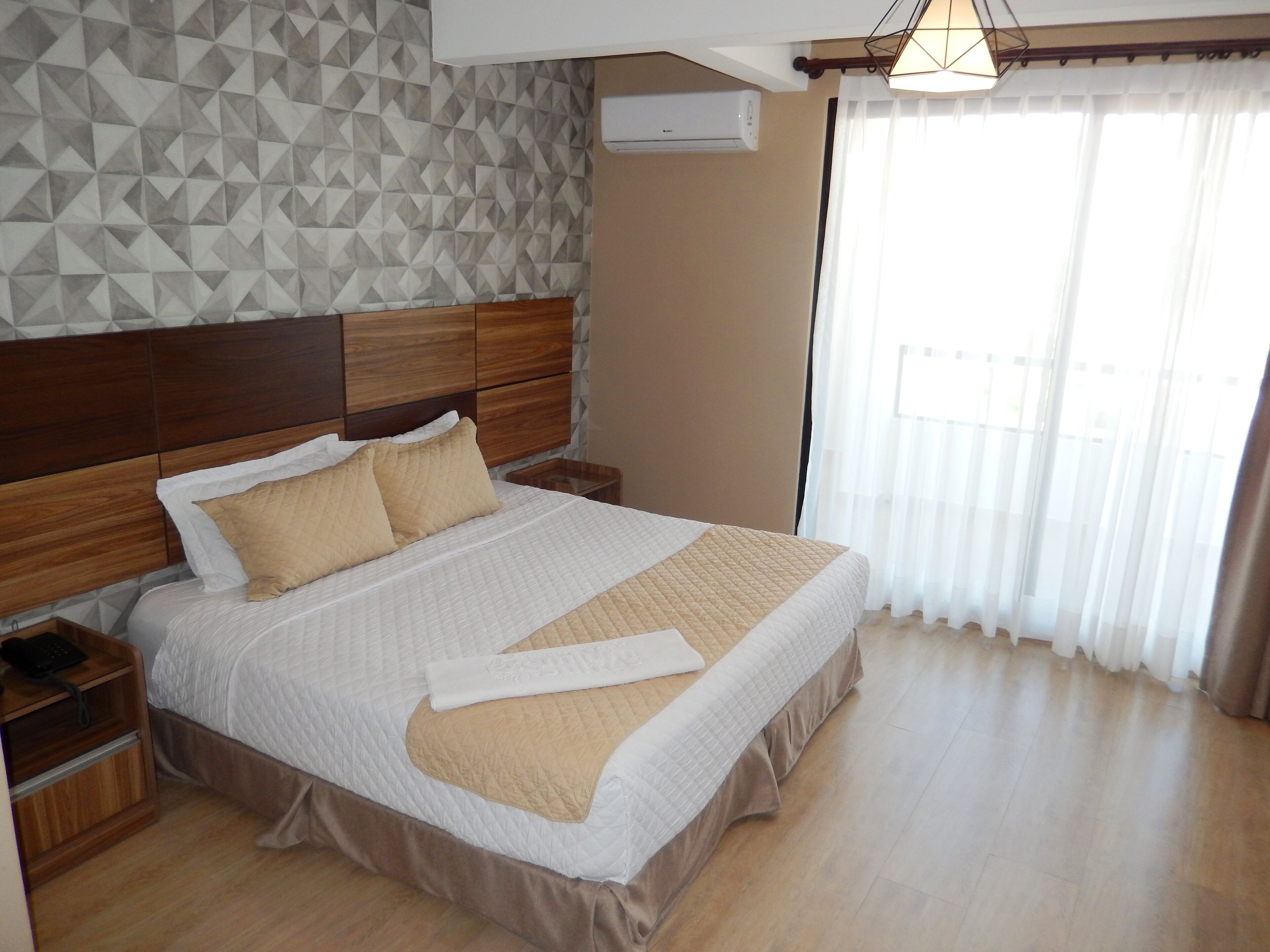 A neatly made bed with a wooden headboard, a bedside table with a phone, and a wall with a geometric pattern.