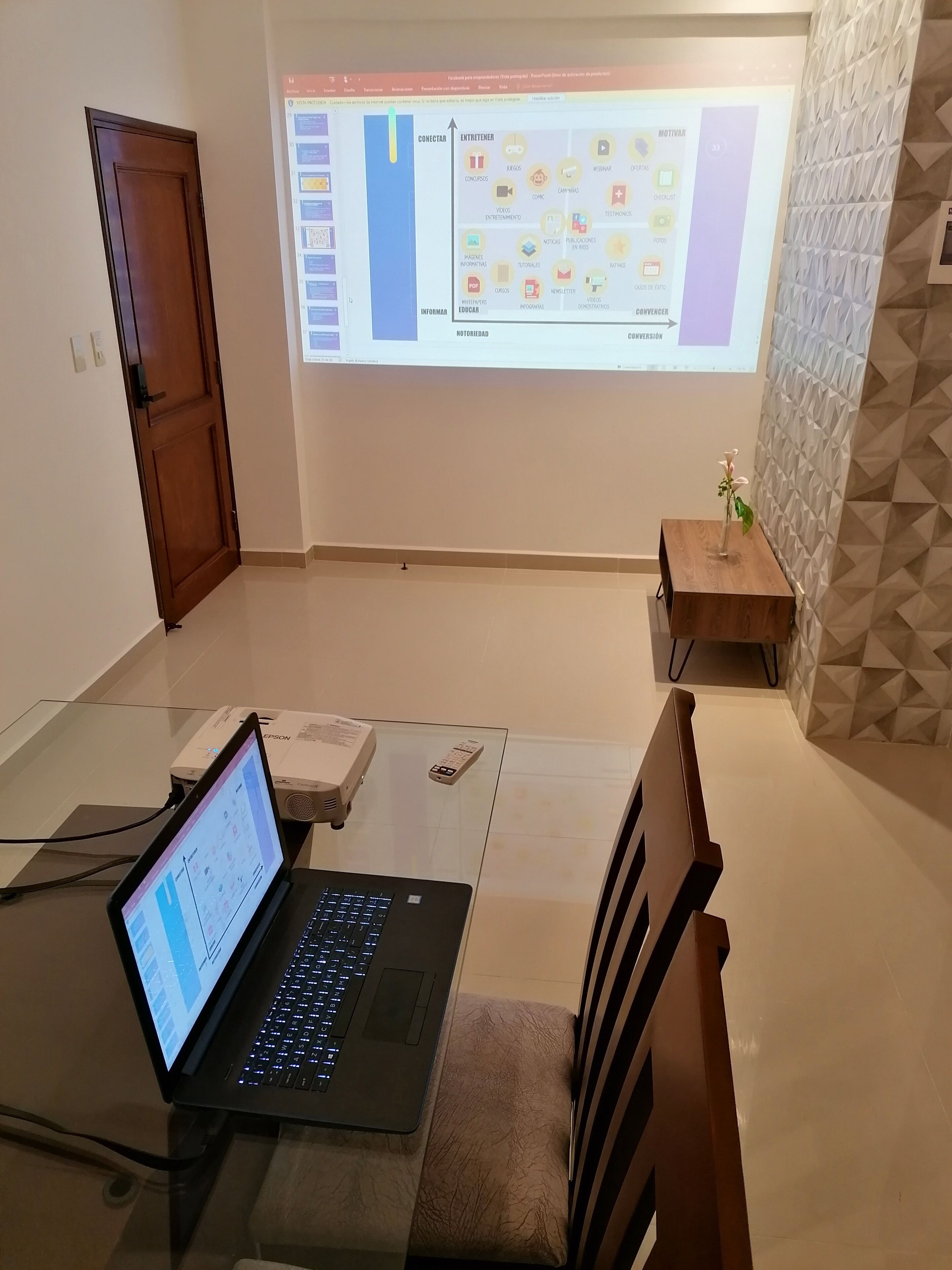 Een kamer met een laptop op een glazen tafel, een beamer die een presentatie toont, een houten deur en een structuurwand.