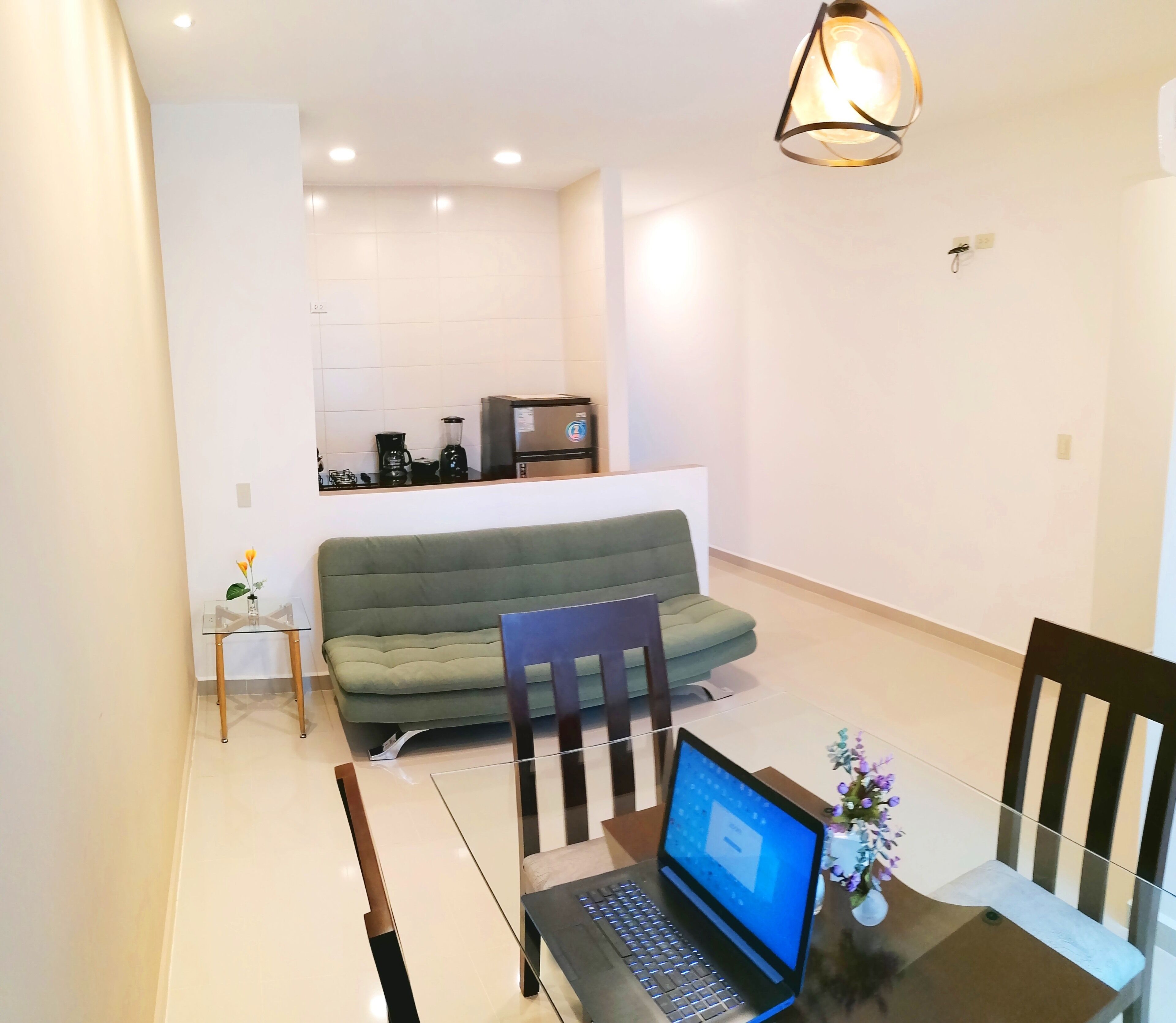 Una sala de estar moderna con un sofá verde, una mesa de comedor con sillas, una computadora portátil y una pequeña mesa con un jarrón de flores.