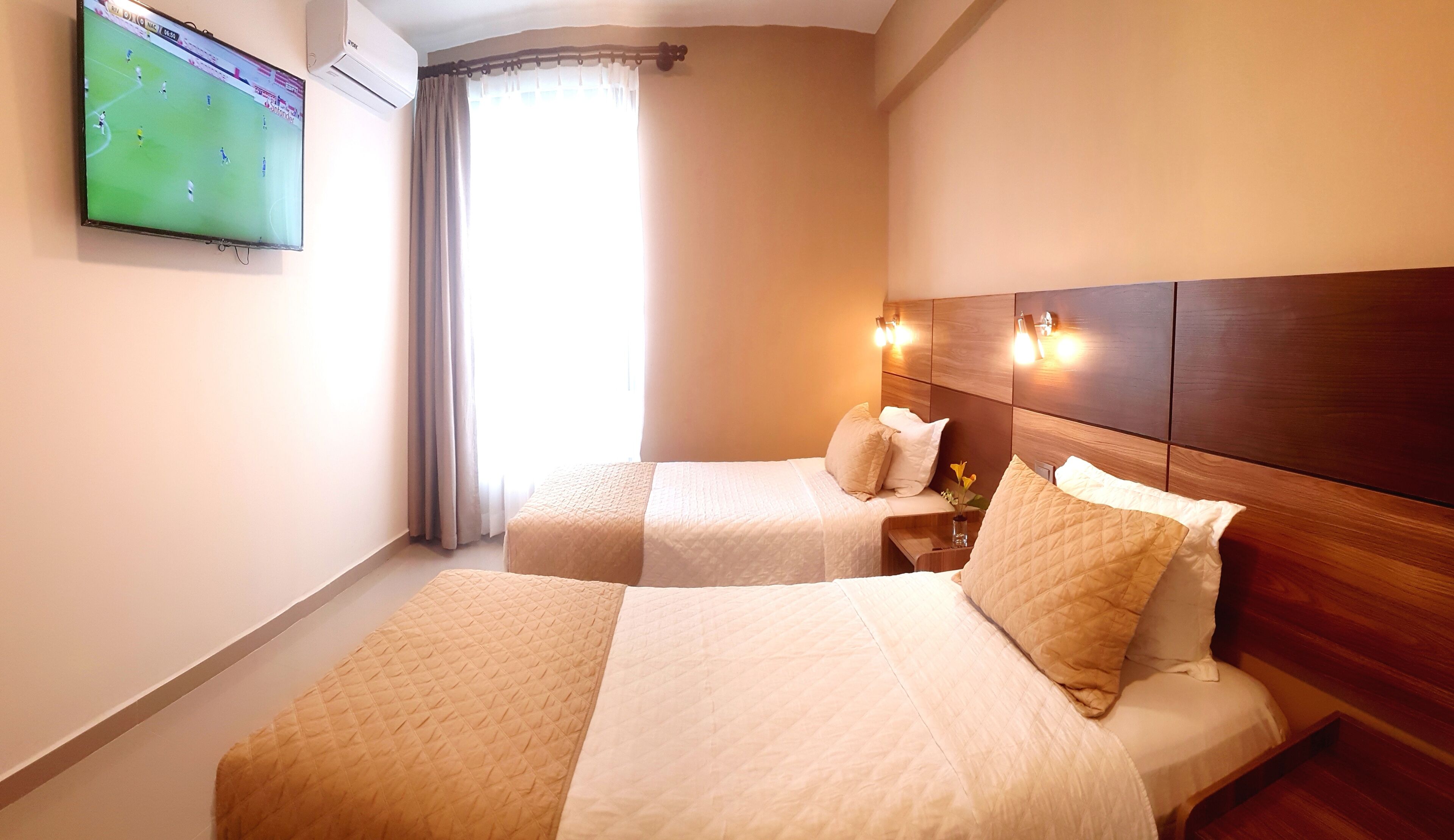 Habitación de hotel con dos camas, un televisor en la pared, una ventana con cortinas y un cabecero de madera con luces de pared.