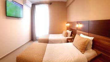 Habitación de hotel con dos camas, un televisor en la pared, una ventana con cortinas y un cabecero de madera con luces de pared.