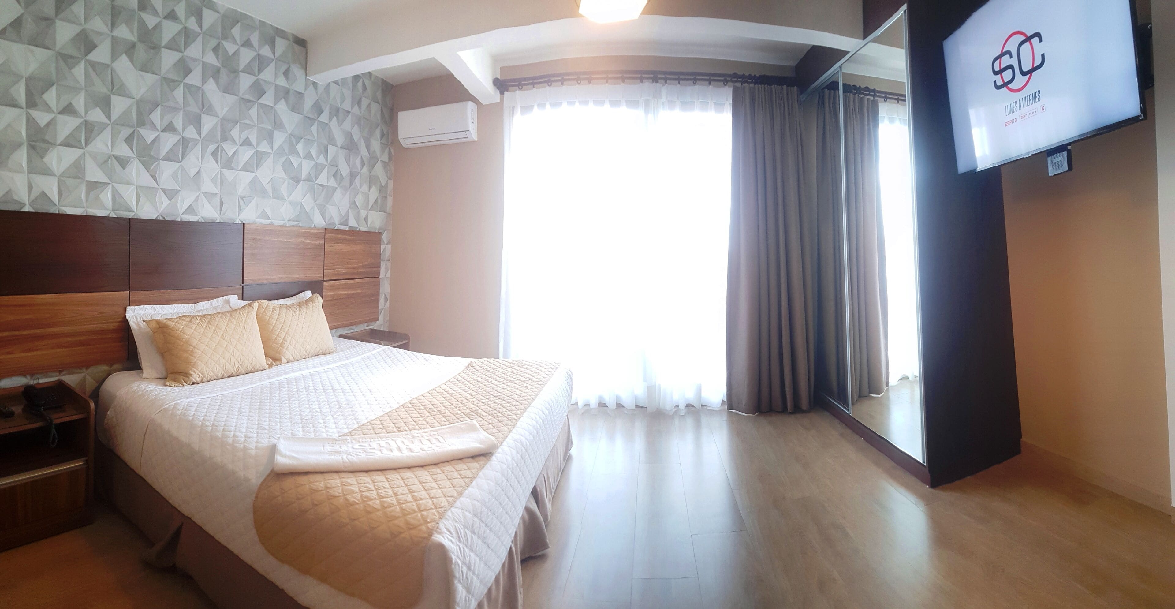 Une chambre d’hôtel avec un grand lit, une tête de lit en bois, un téléviseur à écran plat et une fenêtre avec des rideaux.
