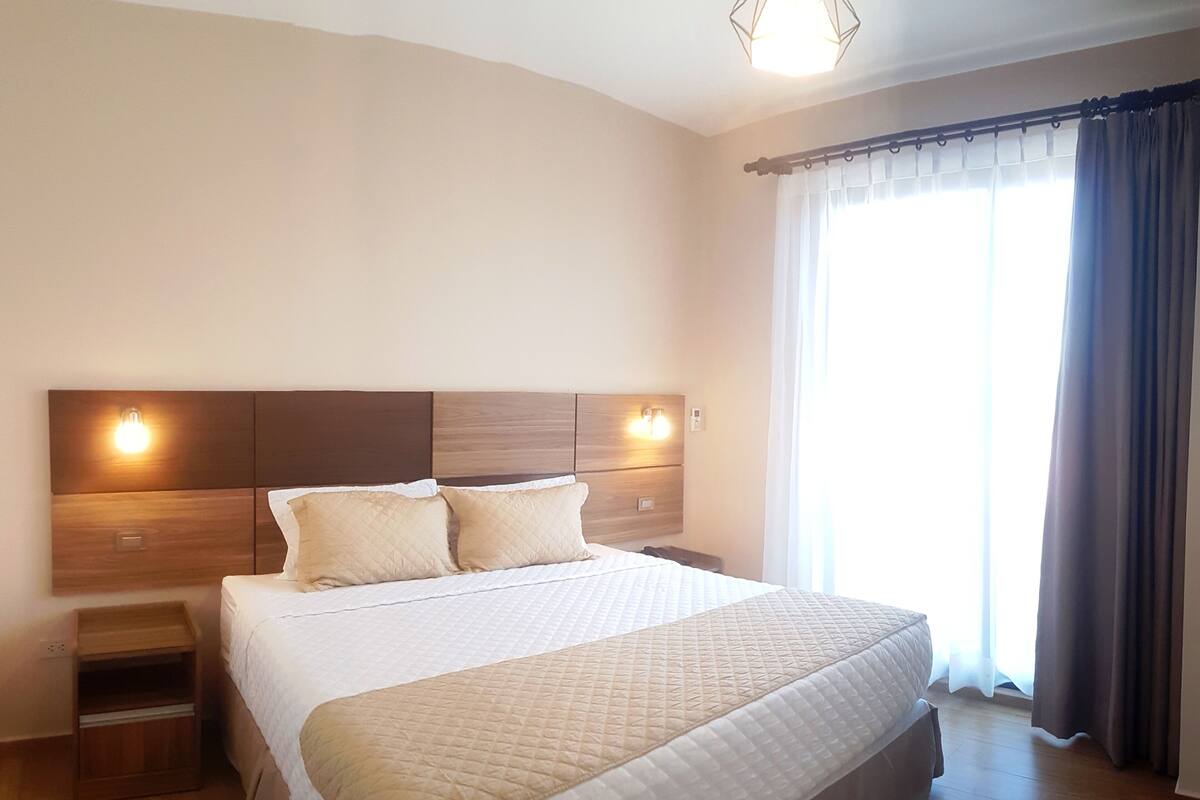 Une chambre d’hôtel moderne dotée d’un grand lit, d’une tête de lit en bois, de tables de chevet et offrant une vue sur l’extérieur grâce à de grandes fenêtres.
