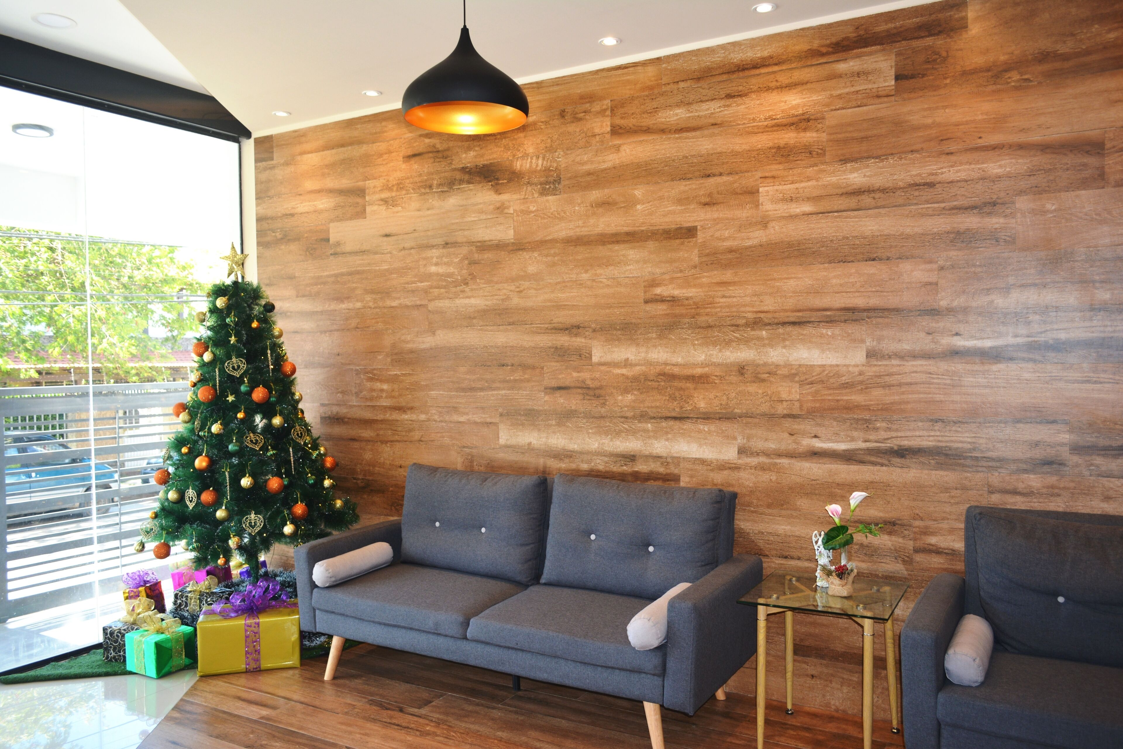 A modern living room with a wooden wall, a grey sofa, a glass coffee table, and a Christmas tree.