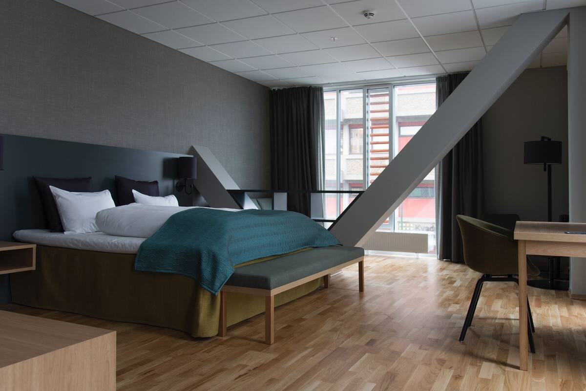 A modern hotel room with a bed, a desk, and a view of the outside.