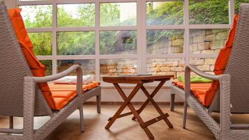 Ein Zimmer mit Korbstühlen und einem Holztisch, große Fenster mit Blick auf Grünflächen und Steinmauern.