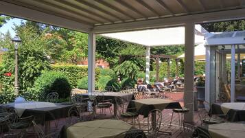 An outdoor patio with tables and chairs, surrounded by greenery and a covered area.