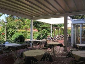 An outdoor patio with tables and chairs, surrounded by greenery and a covered area. - Hotel Am Schlosspark (Gotha)