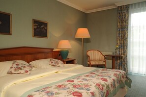 Ein Hotelzimmer mit einem Bett, zwei Lampen, einem Sessel, einem Tisch und einem fensterbepflanzten Fenster.