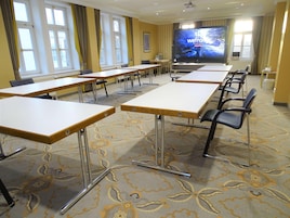Una sala de conferencias con mesas, sillas, una pantalla grande y ventanas con cortinas.