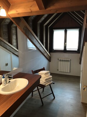 Une salle de bain avec un meuble-lavabo en bois, un lavabo blanc, une chaise avec des serviettes, une fenêtre et un radiateur.