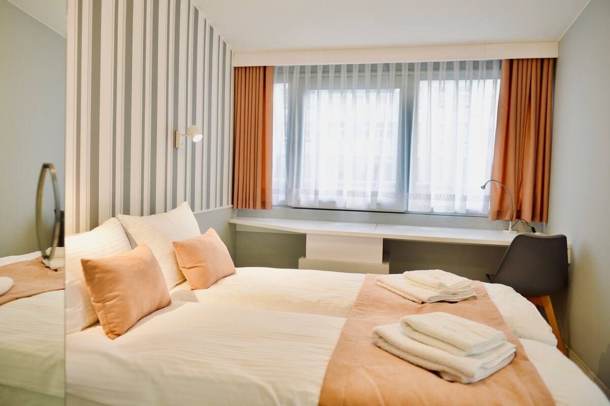 Quarto de hotel com cama, escrivaninha, cadeira e janela com cortinas.