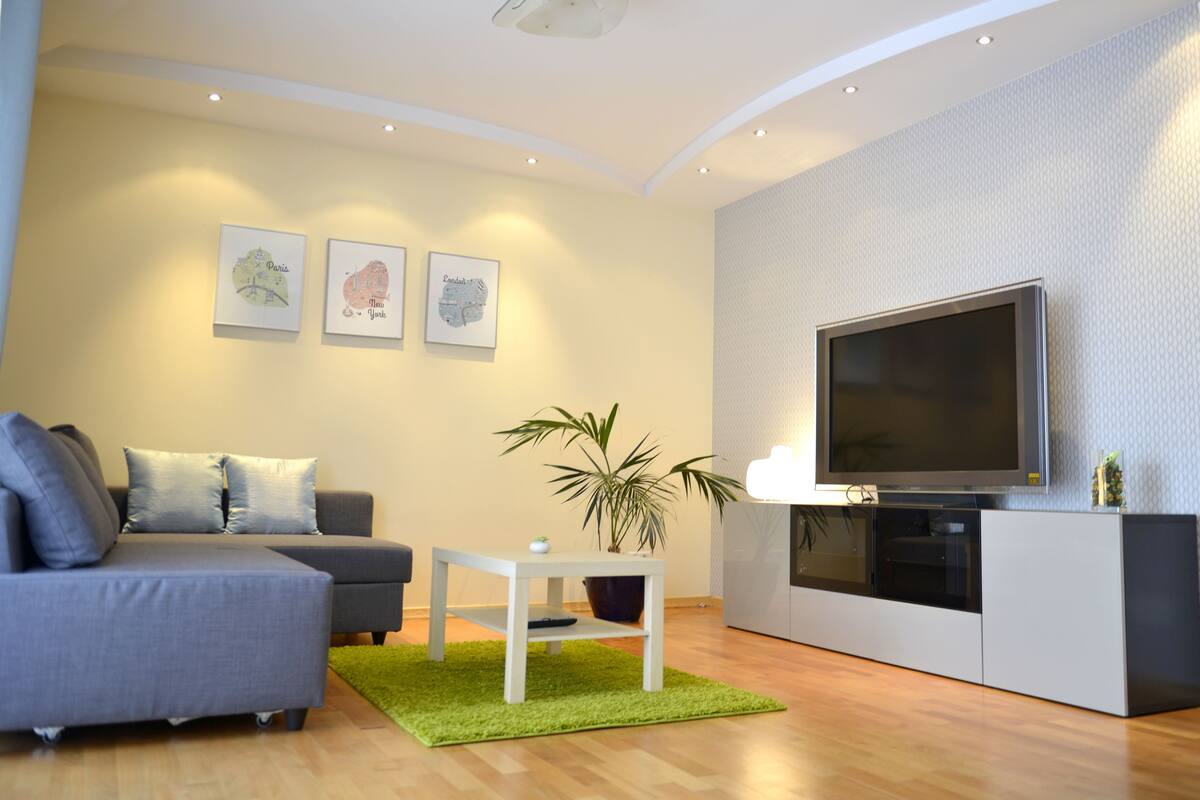 Uma sala de estar moderna com um sofá cinzento, uma mesinha branca, um tapete verde e uma televisão de tela plana sobre um móvel branco.