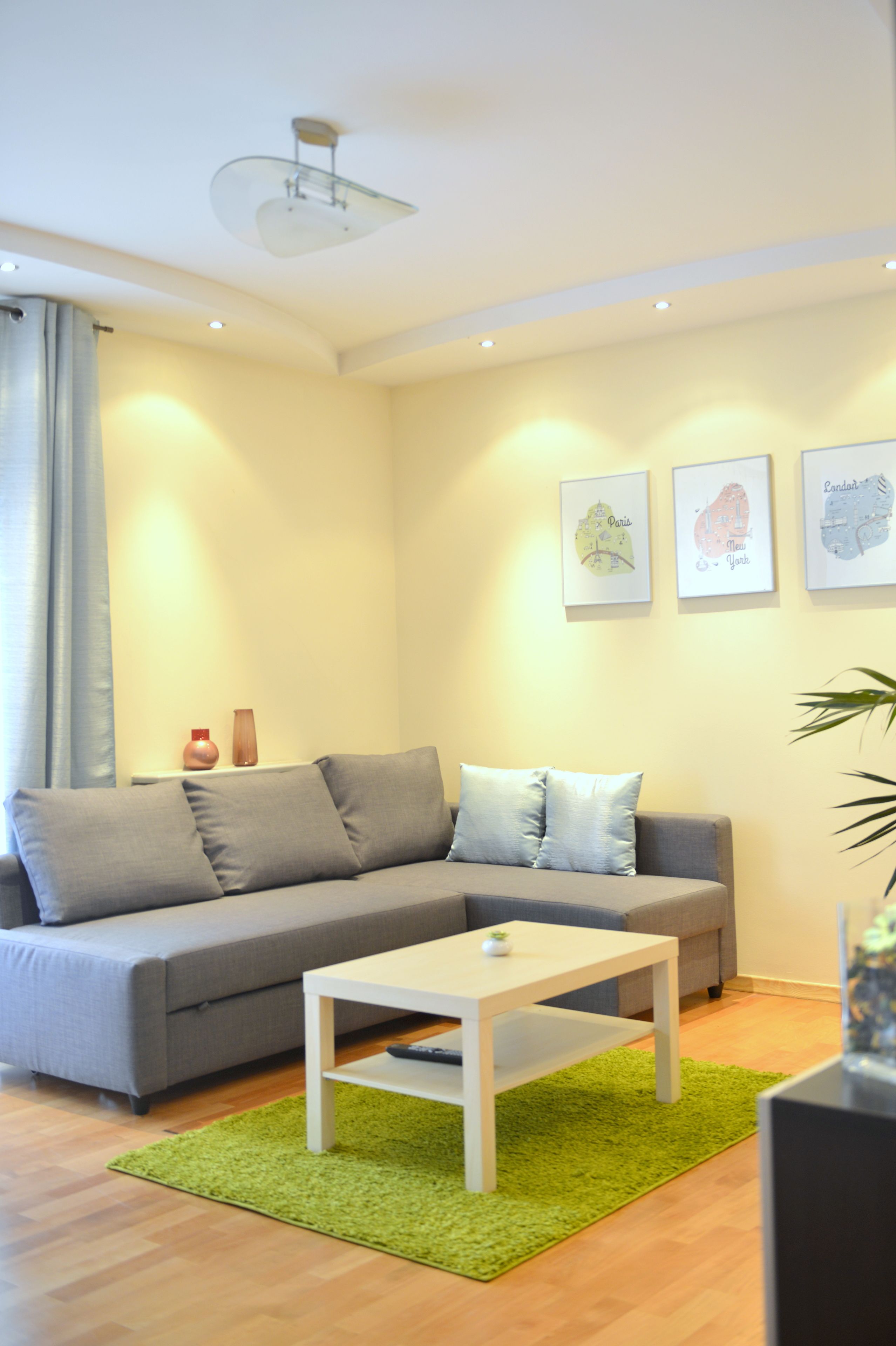 Ein modernes Wohnzimmer mit einem grauen Sofa, einem weißen Couchtisch und einem grünen Teppich. An den Wänden befinden sich Bilder von Stadtansichten in Bilderrahmen.