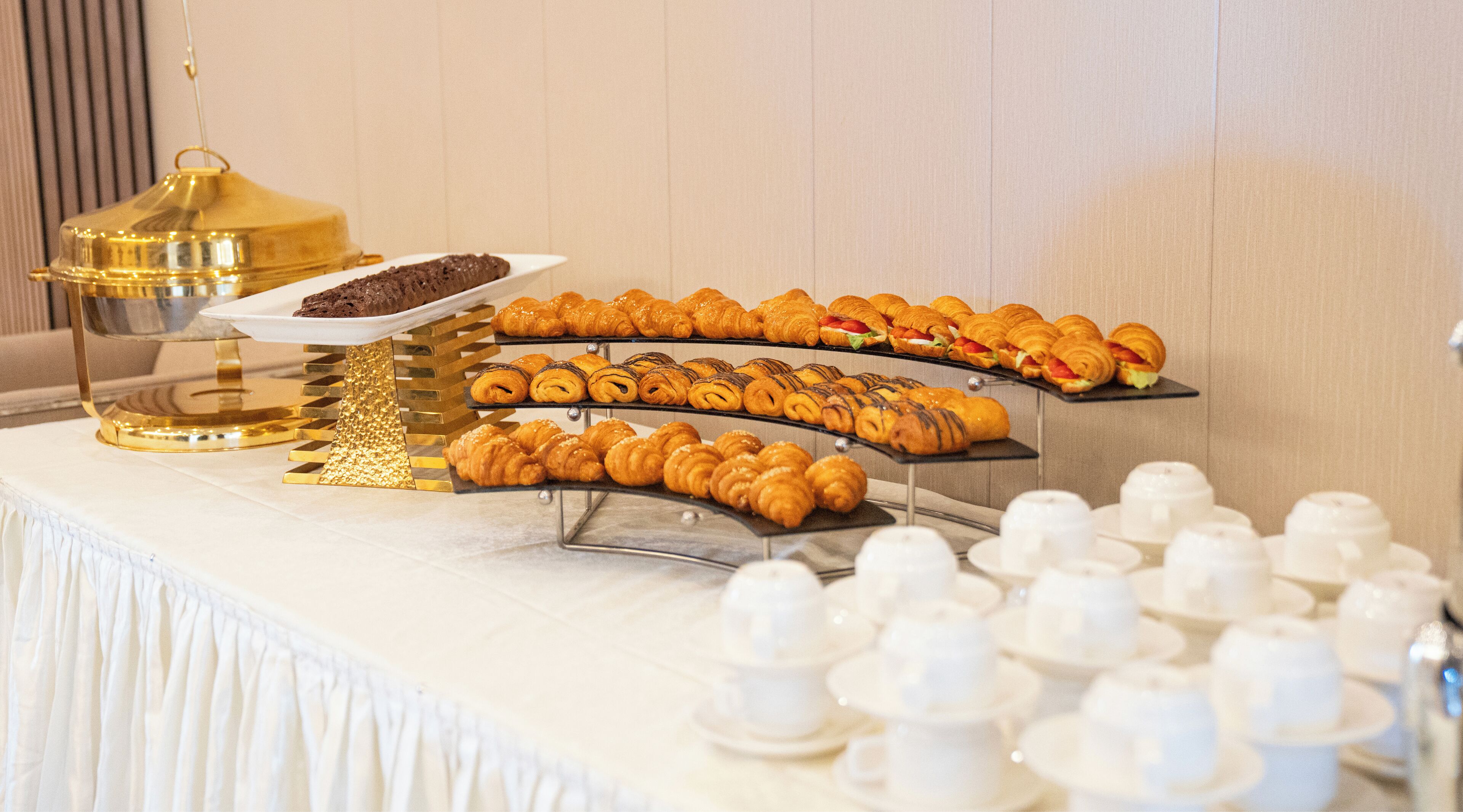 Un bufet ple de pastissos i postres variades, com ara croissants i una torta de xocolata, disposats sobre safates superposades.