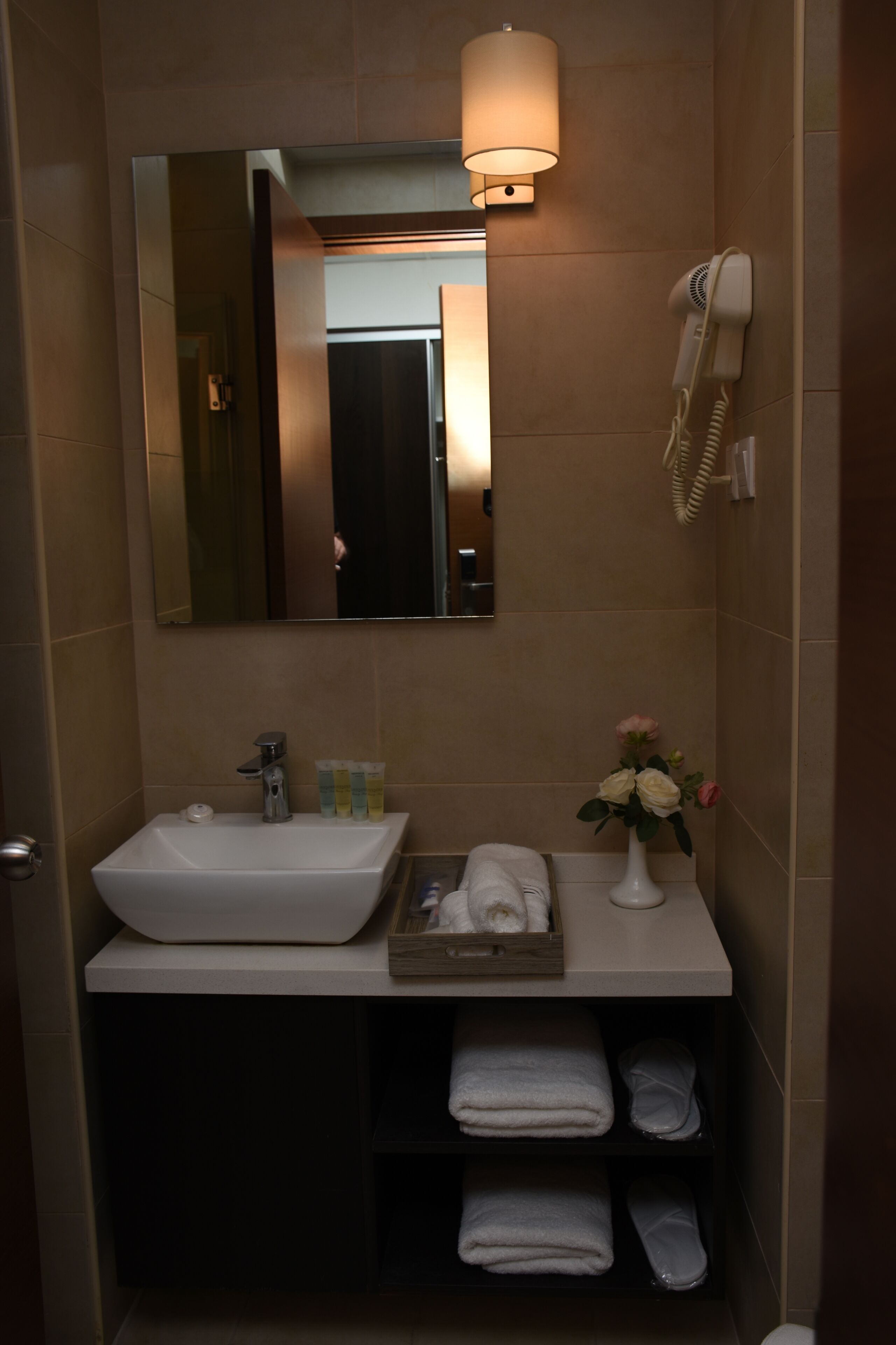 A bathroom with a white sink, a mirror, a hairdryer, and a vase with flowers.