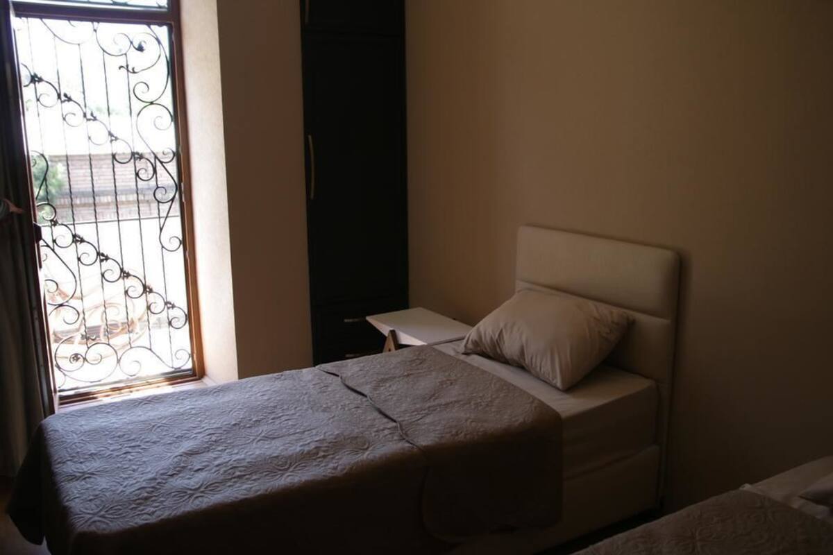 Een enkel bed met hoofdbord, nachtkastje en een raam met decoratieve ijzeren spijlen.