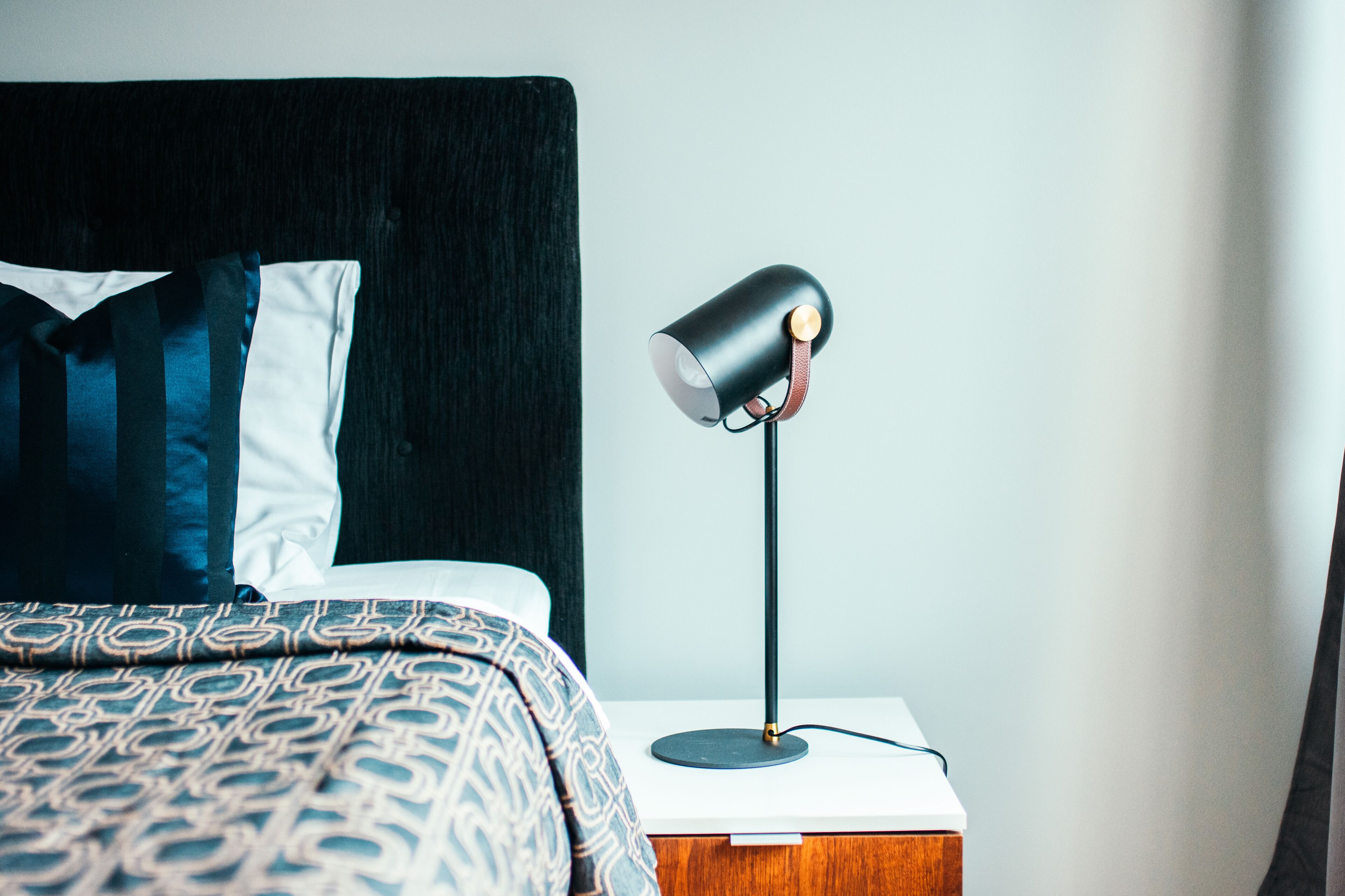 Cama bem arrumada com roupa de cama estampada, cabeceira preta e mesa de cabeceira com luminária moderna.