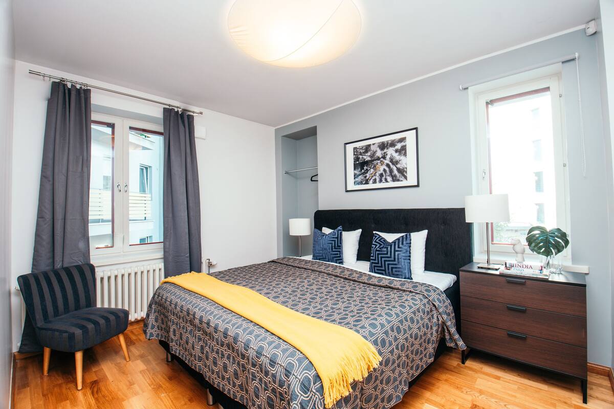 A bedroom with a bed, a chair, a bedside table, a lamp, and a window.