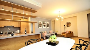 Cozinha moderna com uma ilha central, sala de jantar com mesa branca e cadeiras pretas, e sala de estar com sofá e mesa de centro redonda.