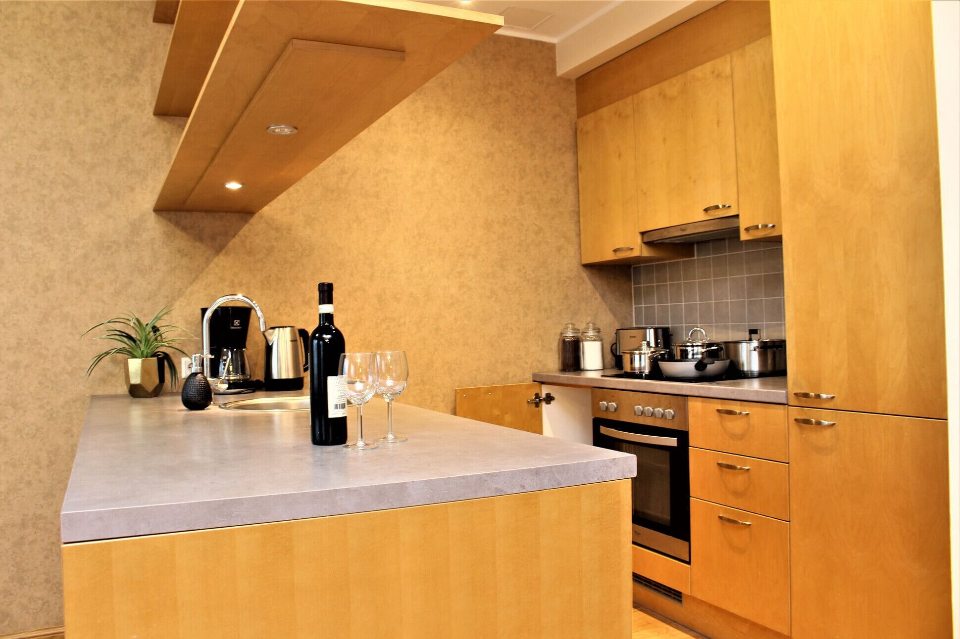 Een moderne keuken met houten kastjes, een inbouwoven en een aanrecht met een koffiezetapparaat, een fles wijn en glazen.