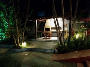 A nighttime outdoor area with a covered seating area, a stone structure, and surrounding vegetation. - Pousada Olhar da Sereia (Porto Seguro)