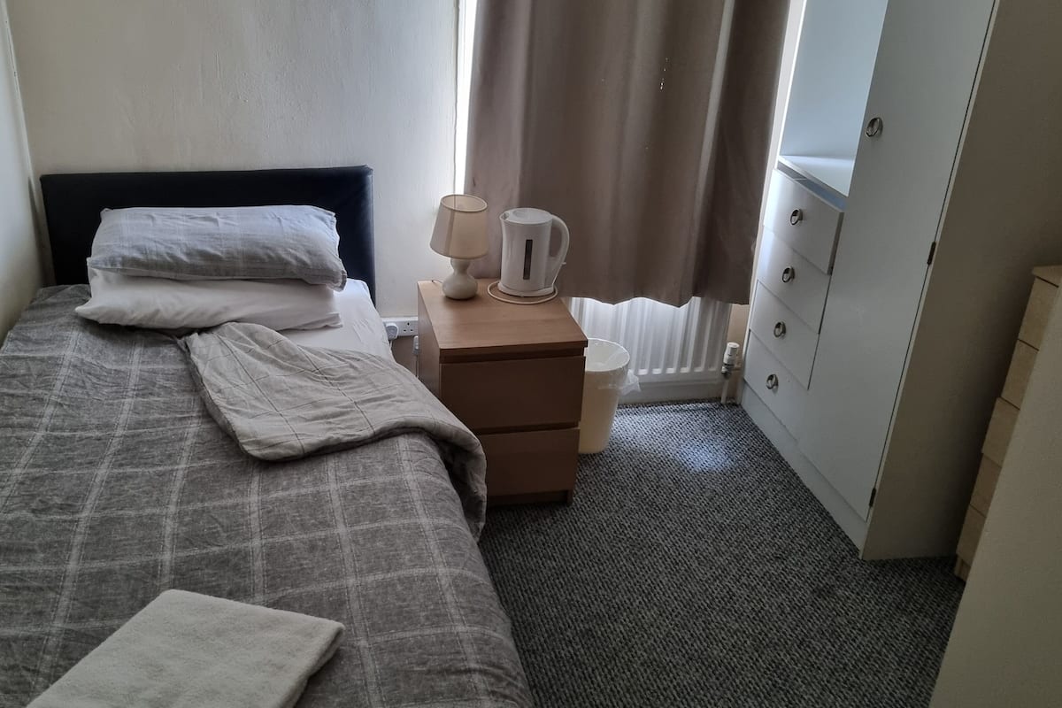 A small, neatly made bed with a grey checkered bedspread and white pillows, a bedside table with a lamp and a white pitcher, and a radiator next to a door.