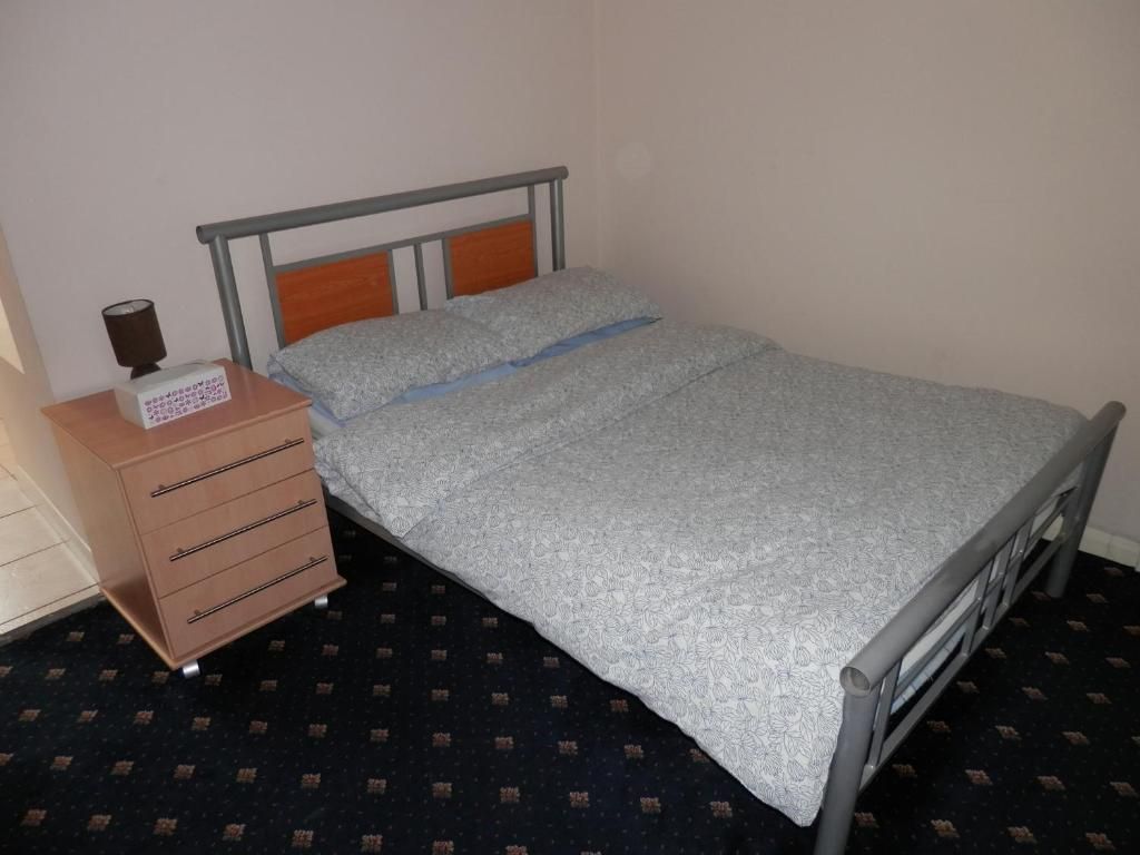 Uma cama de solteiro com estrutura de metal, cabeceira de madeira e mesa de cabeceira com gavetas e abajur.
