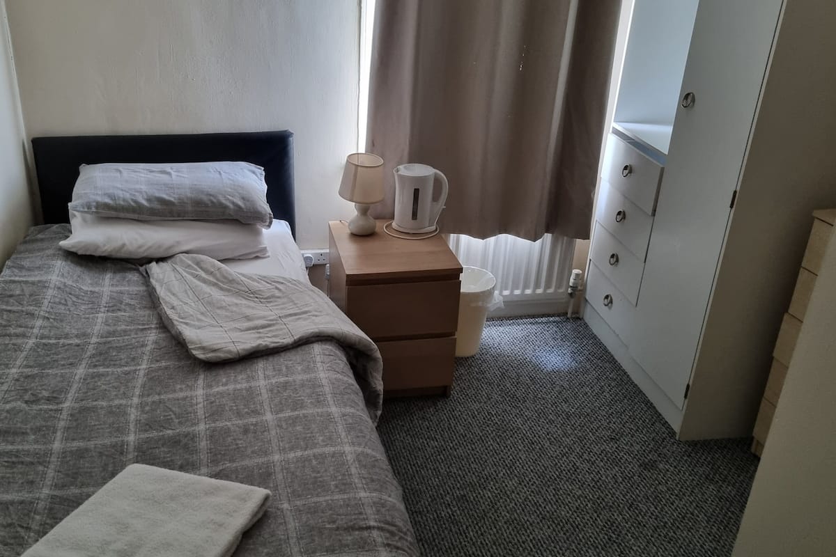 A small, neatly made bed with a grey checkered bedspread and pillows, a bedside table with a lamp and a kettle, a radiator, and a wardrobe with a mirror.