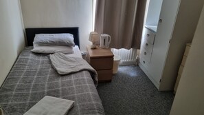 A small, neatly made bed with a grey checkered bedspread and pillows, a bedside table with a lamp and a kettle, a radiator, and a wardrobe with a mirror. - Shamrock Guest House (Gravesend)