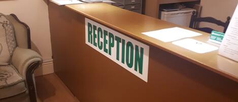 A reception area with a sign that reads "RECEPTION" on a brown counter. There is a chair to the left and a television mounted on the wall above the counter.