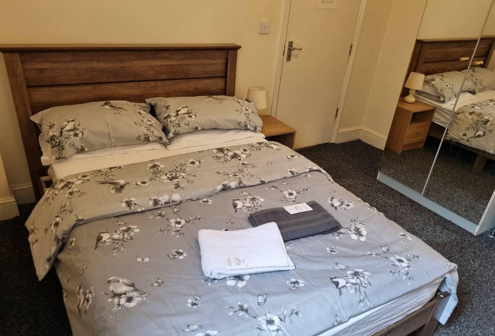 A neatly made bed with floral bedding, a bedside lamp, and a mirror on the wall.