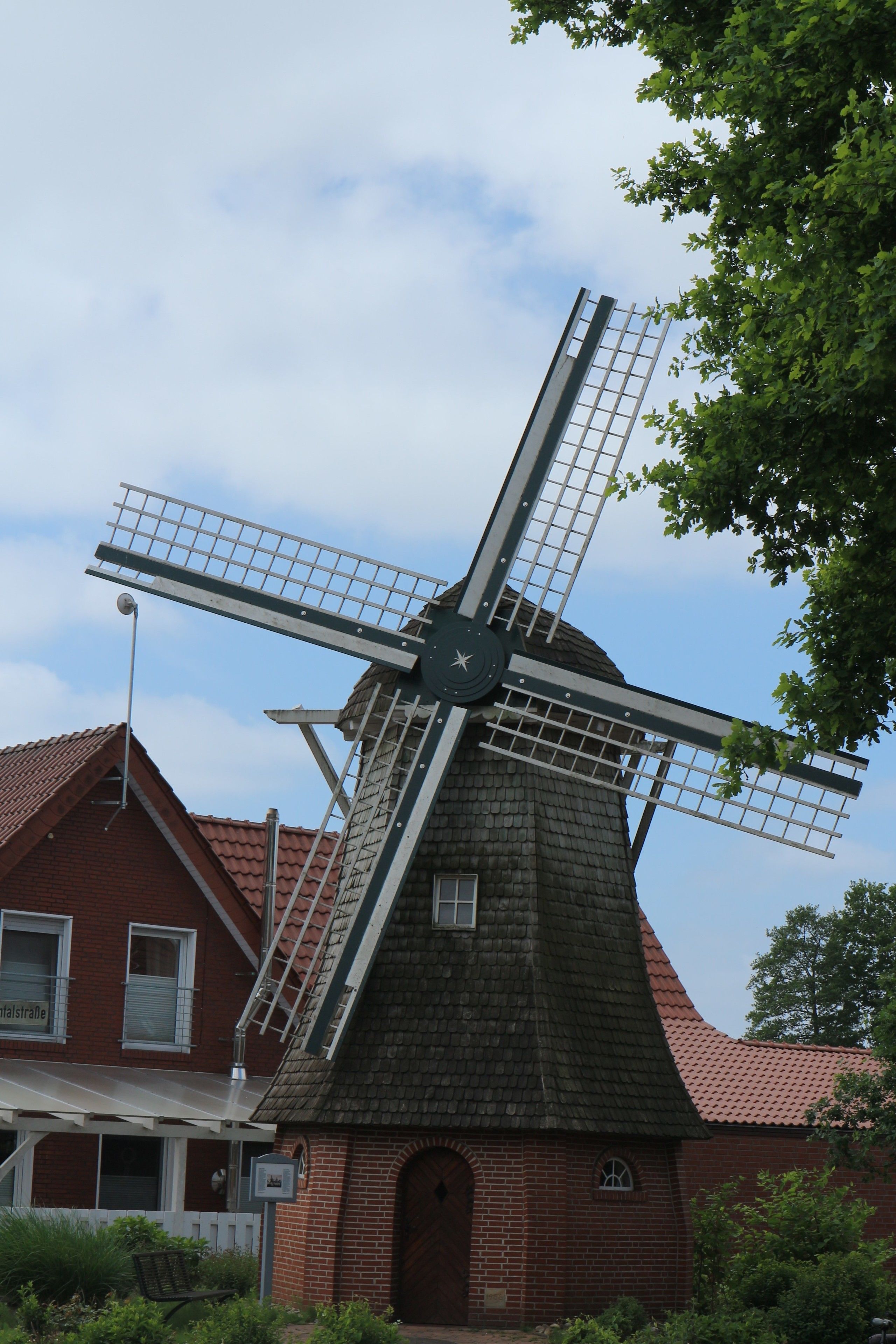 A traditional windmill with a brick base and a sloped roof, adjacent to a red-brick building with a white porch and a visible window.