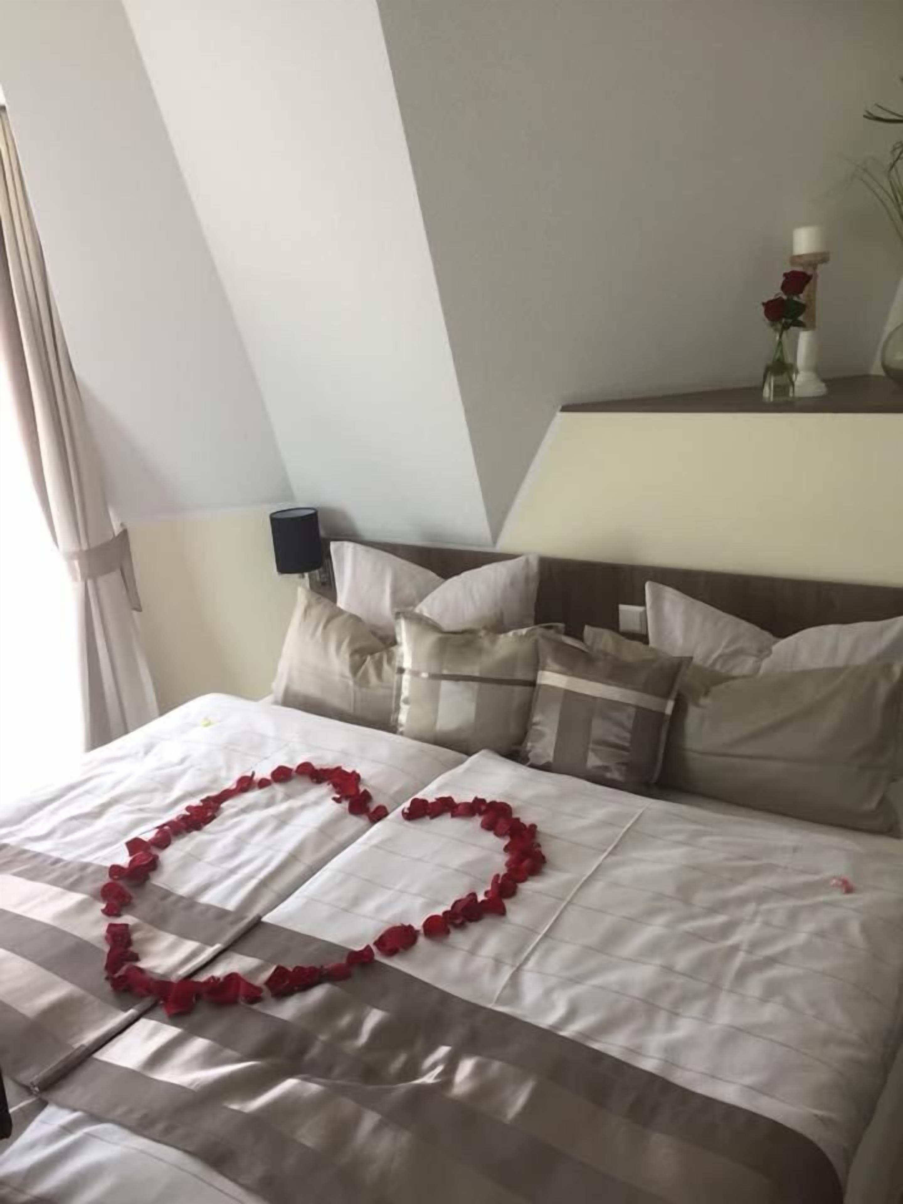 Une chambre d’hôtel avec un lit parfaitement fait, une composition en forme de cœur faite de pétales de roses et un vase contenant une seule rose rouge sur la table de chevet.