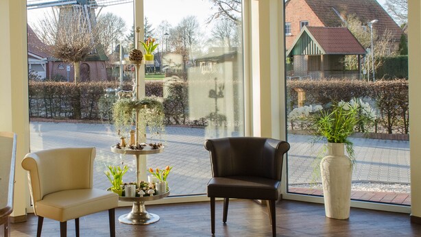 A room with a large window, a black chair, a white chair, and a small table with plants. - Gasthof Robben (Twist)