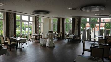 A spacious dining area with multiple tables and chairs, large windows offering views of the outside, and a ceiling with decorative elements.