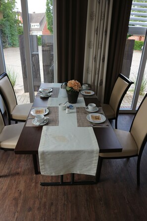 Una mesa de comedor con platos, tazas y cubiertos, rodeada de sillas, con vista a una zona residencial a través de grandes ventanales.