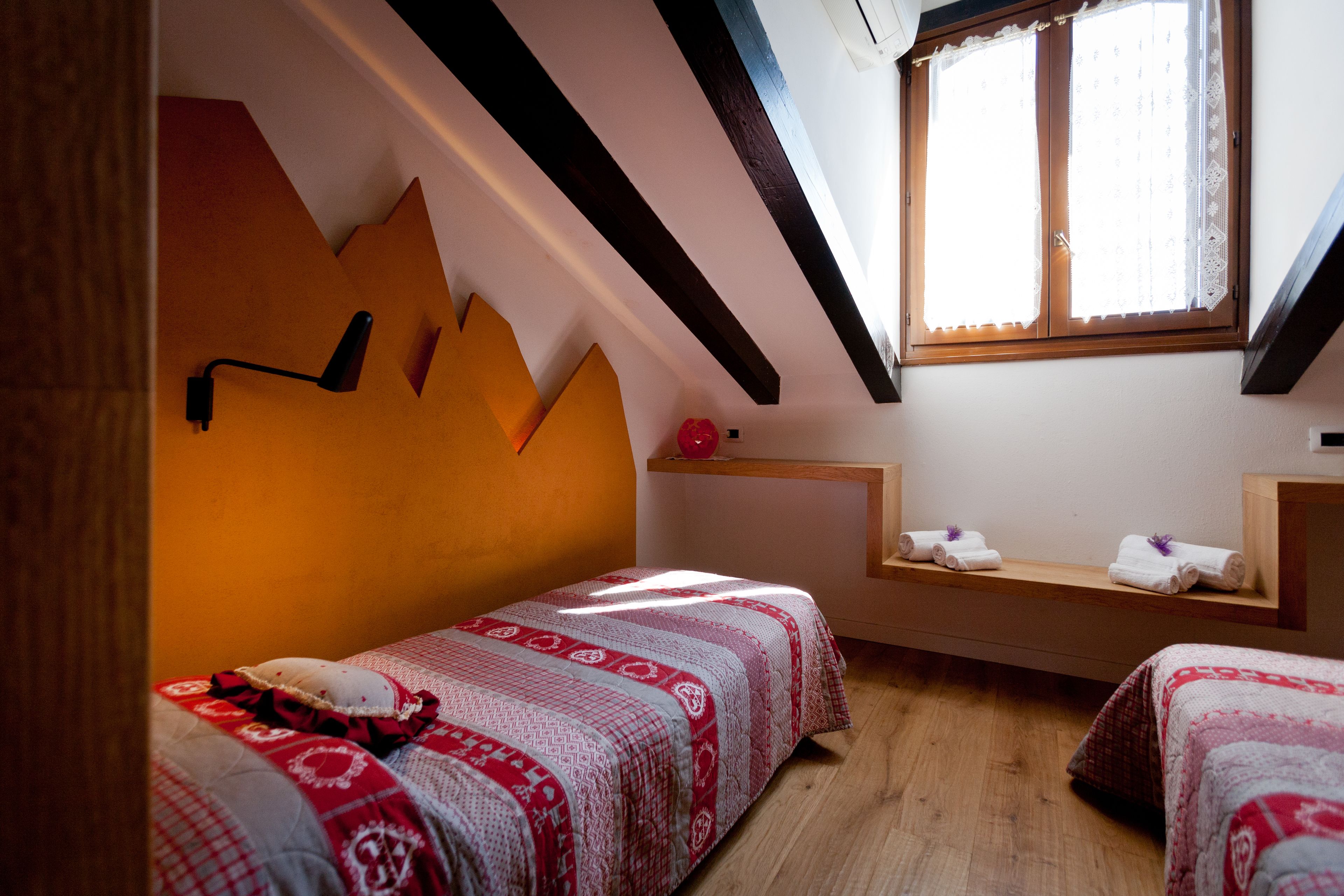 A bedroom with two beds, a window with lace curtains, a wooden ceiling, and a small shelf with towels.