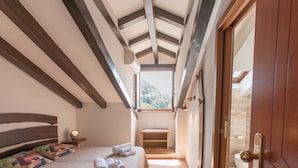 A bedroom with a sloped ceiling, a bed with a wooden headboard, a bedside table with a lamp, and a view of a bathroom through an open door. - Grand Hotel Gortani Wellness & Relax (Arta Terme)