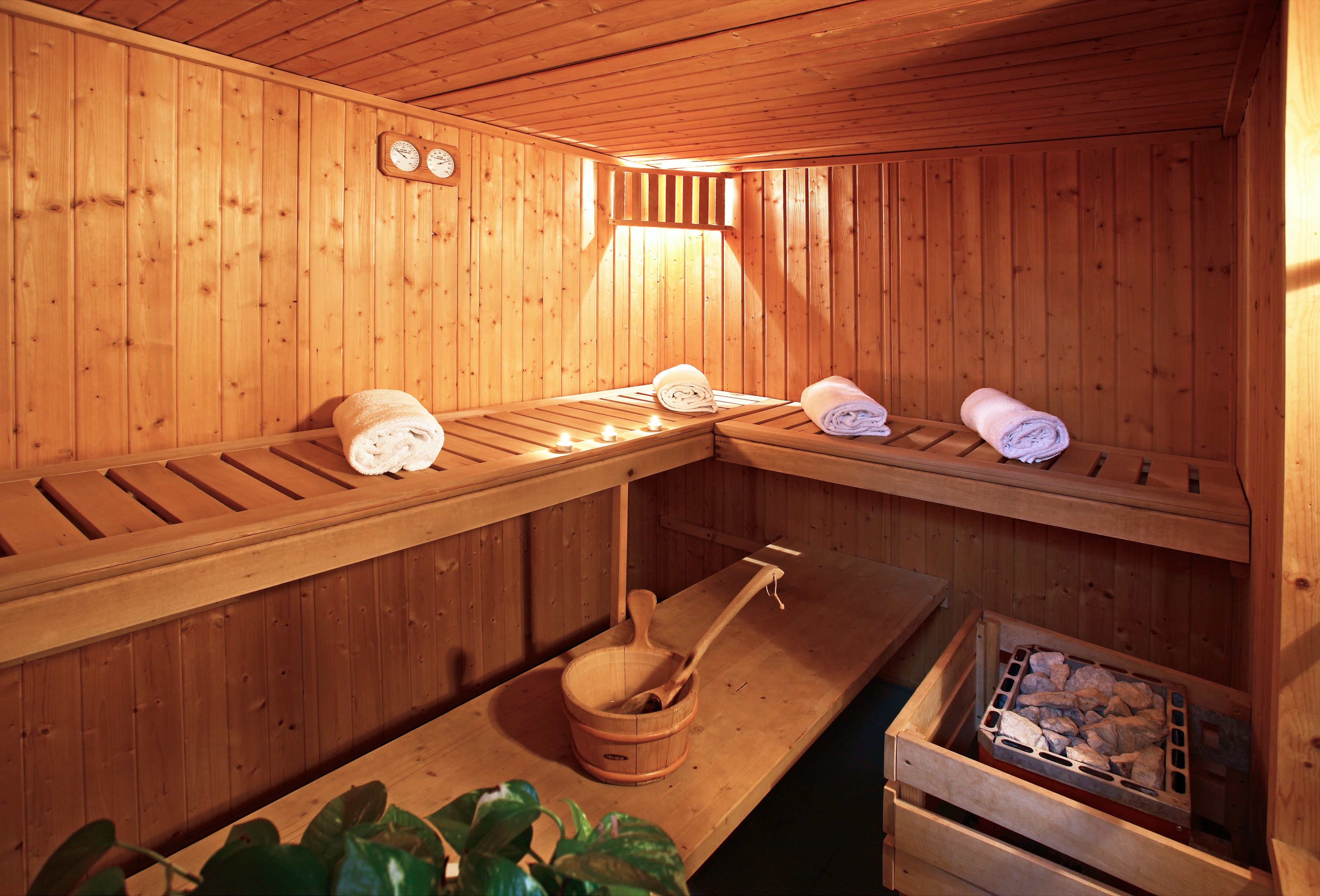 Una sauna con paredes de madera, un banco, un banco con toallas, un recipiente de madera y un contenedor lleno de piedras.
