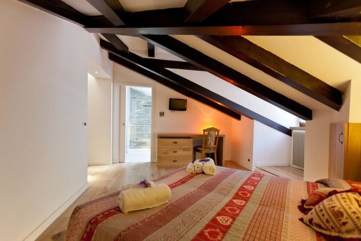 Une chambre avec un plafond en pente, un lit recouvert d’une couvre-lit à motifs, une chaise, un bureau avec un ordinateur et une fenêtre donnant sur l’extérieur.