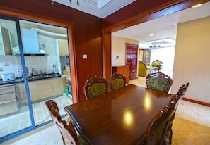 A dining area with a wooden table and chairs, a kitchen in the background, and a chandelier in the dining room.