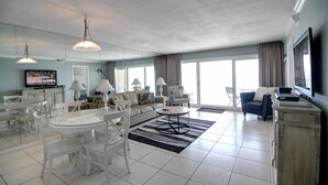 A modern living room with a dining area, large windows, and a flat-screen TV. - Beach House Condos by Crystal Waters (Destin)