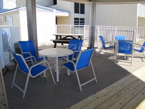 A covered outdoor seating area with a round table and blue chairs, wooden deck flooring, and a white railing. - Beach House Condos by Crystal Waters (Destin)