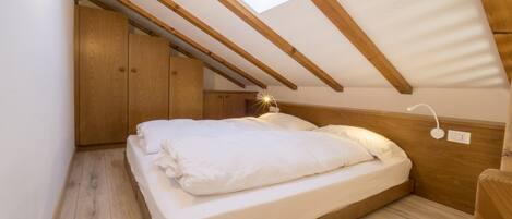 A bedroom with a sloped ceiling, wooden beams, a skylight, a bed with white bedding, and a wooden wardrobe.