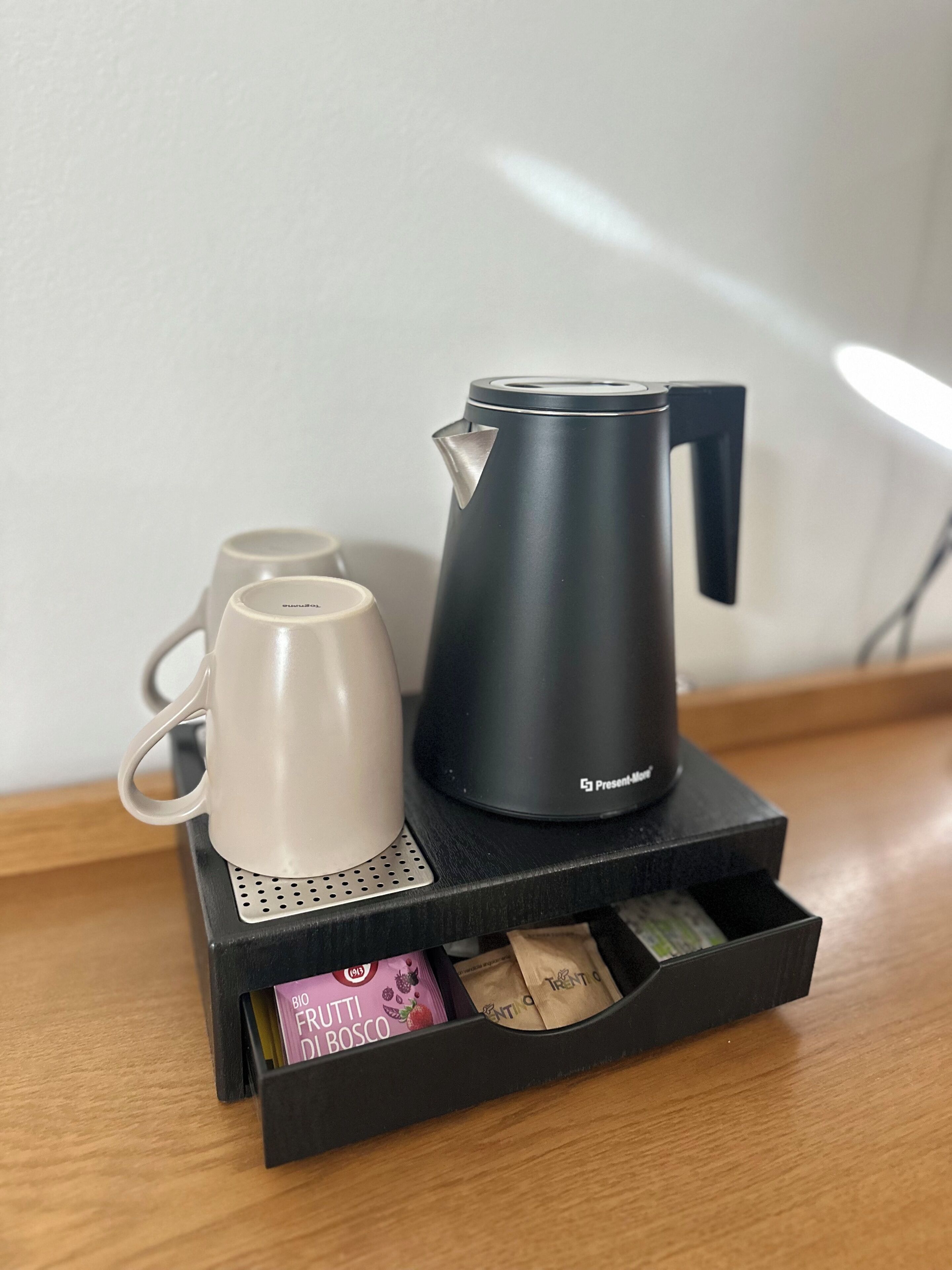 A black coffee maker on a tray with two white cups and a drawer containing tea bags.
