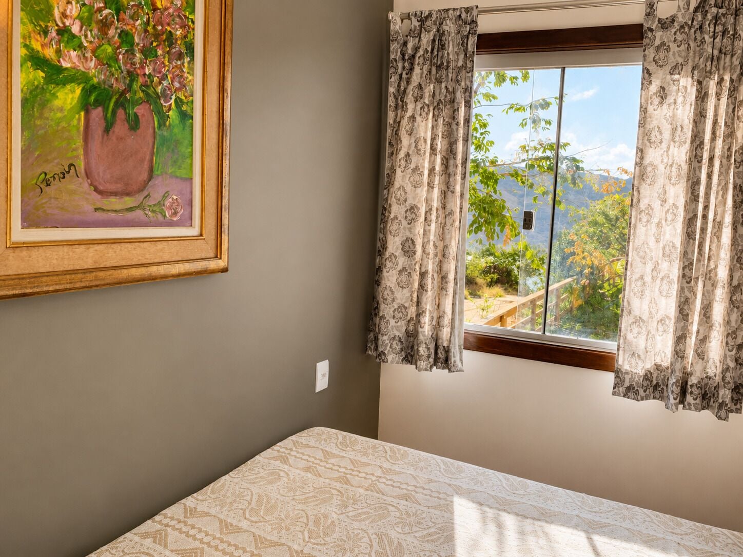A room with a bed, a window with curtains, a painting on the wall, and a view of greenery outside.
