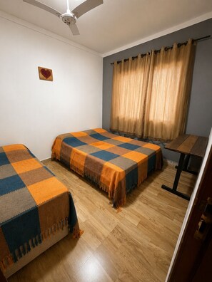 Una habitación con dos camas, un ventilador de techo, una ventana con cortinas y una mesita.