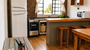 A kitchen with a refrigerator, stove, and microwave. There is a wooden table and stools, and a window with curtains. - Charme e Aconchego - Casa para 10 pessoas com 3 quartos (Capitólio)